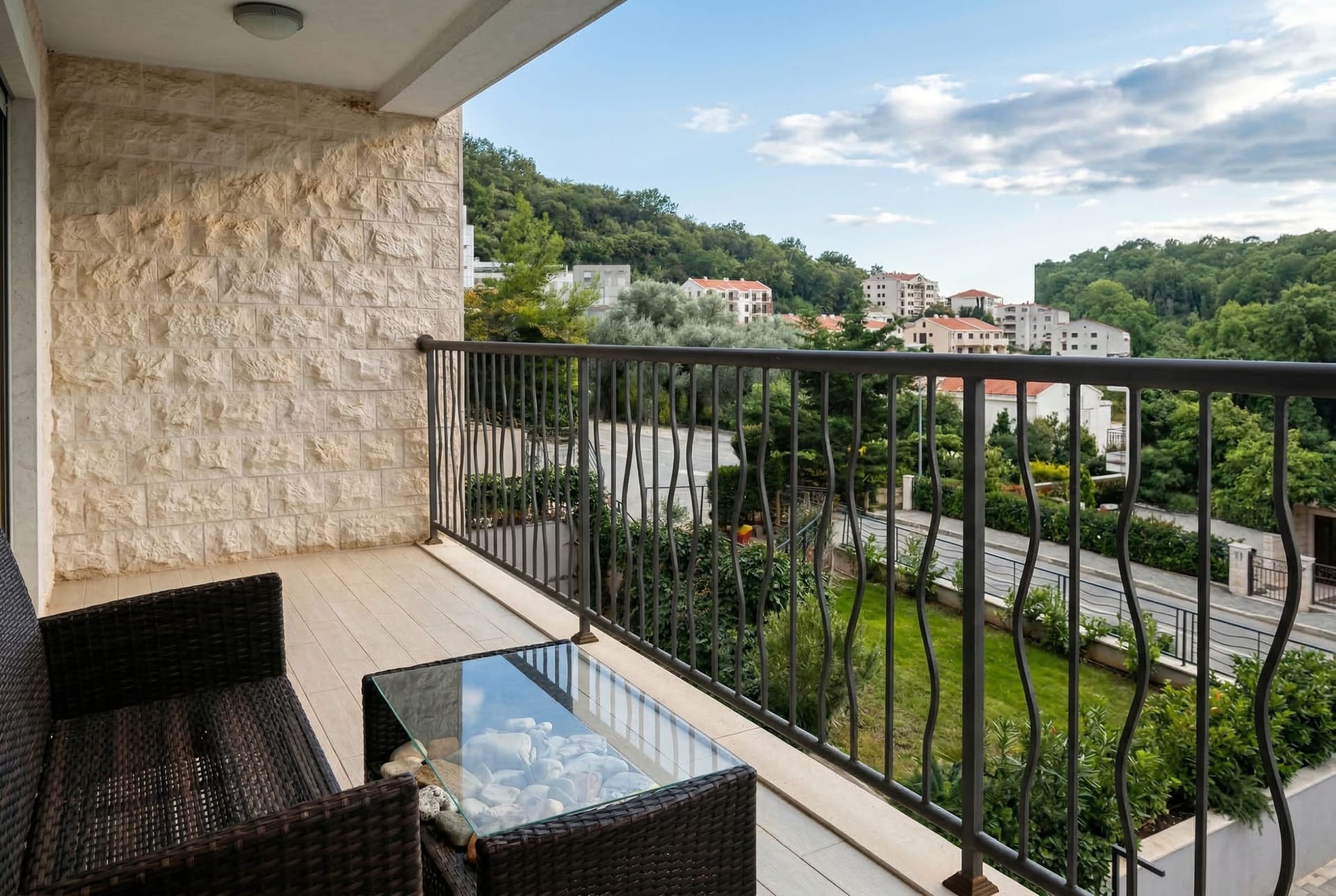 Balcony with wicker chairs and a stone-accent wall in an apartment in Pržno, Montenegro, overlooking green hills.