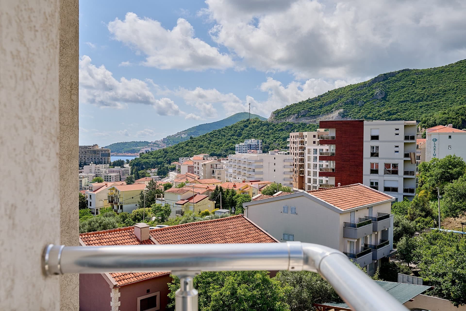 Pogled sa balkona stana u Bečićima, Crna Gora: panorama grada, zelene planine i morska obala u daljini.