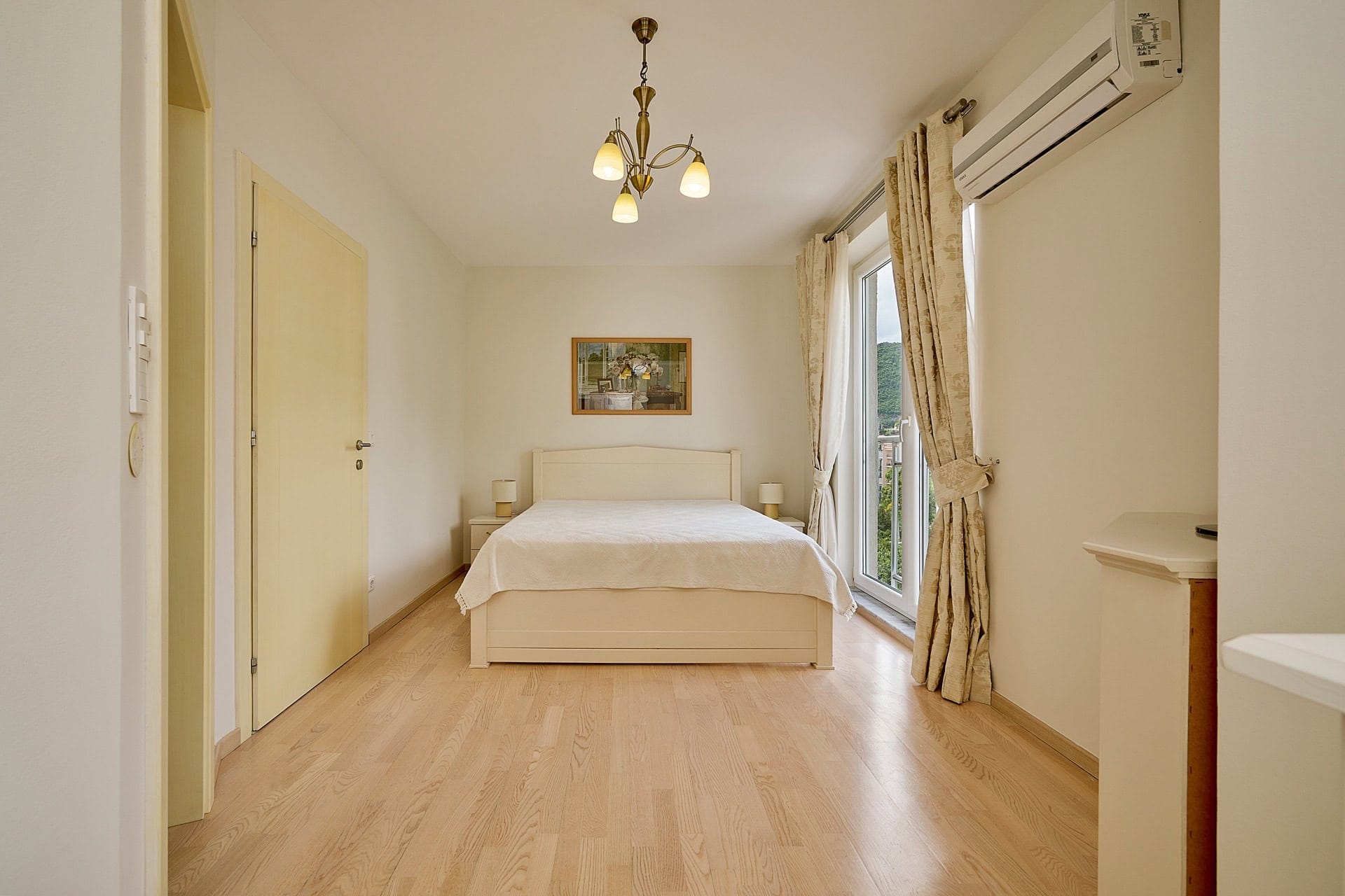 Svetla spavaća soba sa bračnim krevetom u stanu u Bečićima, Crna Gora: parket, klima-uređaj i izlaz na balkon.