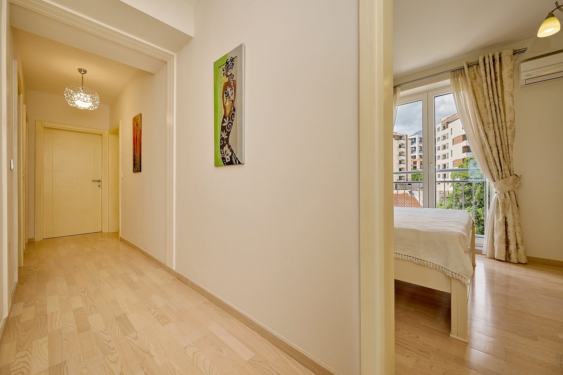 Svetao hodnik i spavaća soba sa izlazom na balkon u stanu u Bečićima, Crna Gora: parket, dekor i pogled na grad.