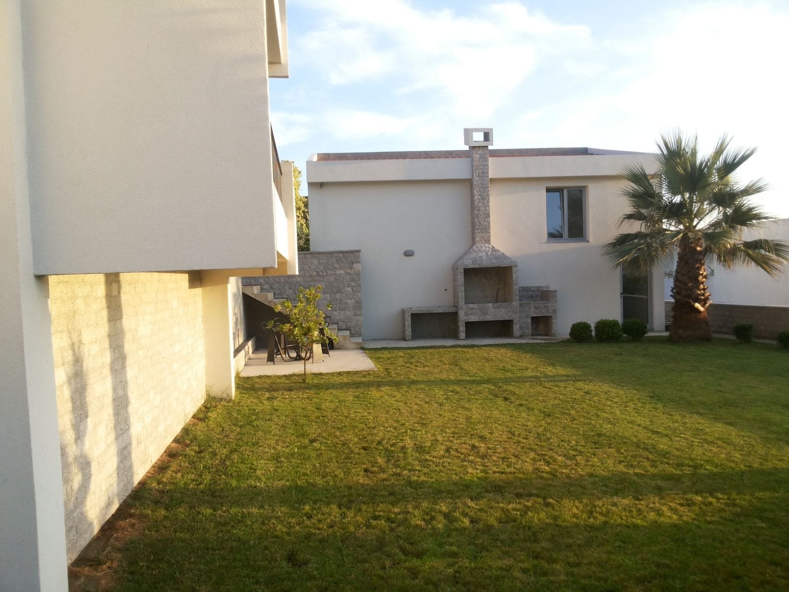 Dvorište privatne kuće u Baru, Crna Gora: zelena travnata površina, kameni roštilj, palma i zona za odmor uz zid zgrade.