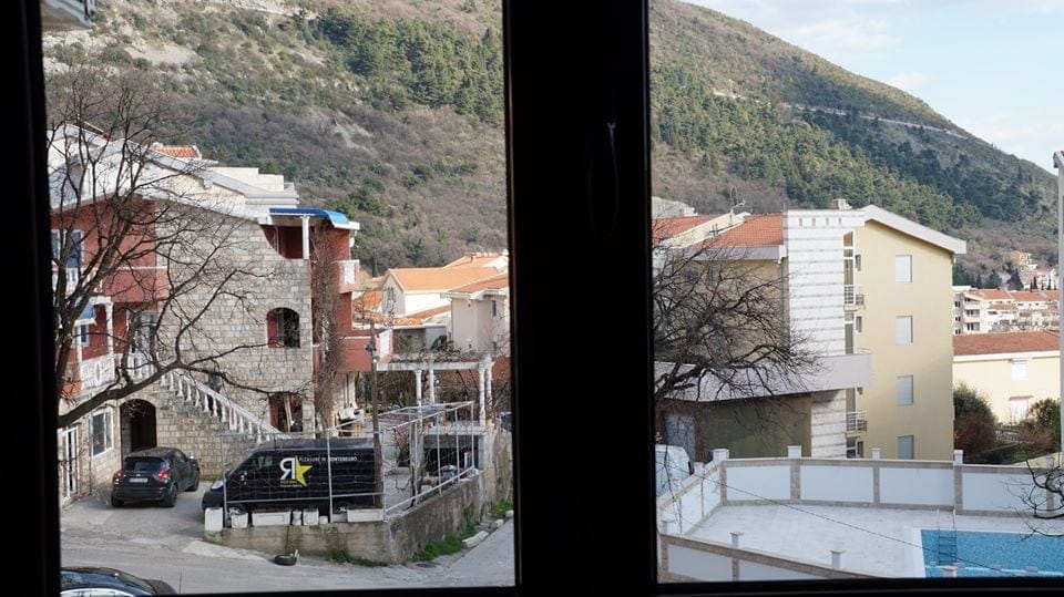 Pogled iz prozora stana u Budvi, Crna Gora: panorama na planine, stambene zgrade i bazen u dvorištu stambenog kompleksa.