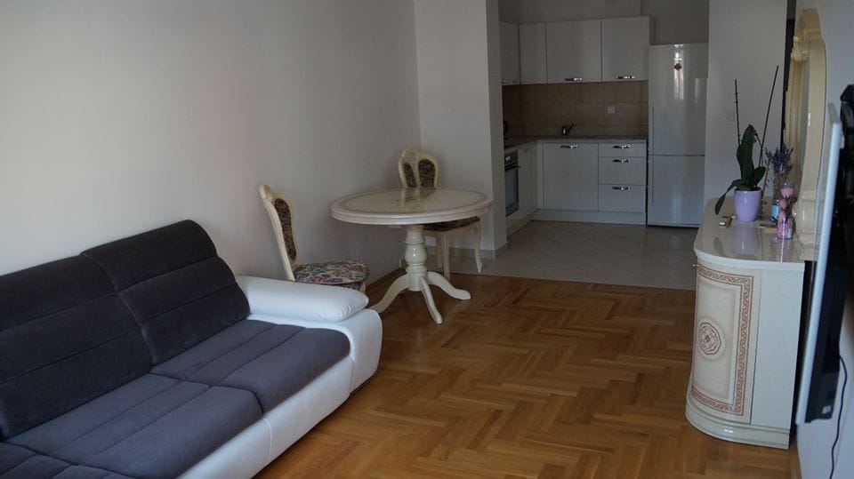 Dnevna soba sa kuhinjom u stanu u Budvi, Crna Gora: sofa, trpezarijski sto, svetao nameštaj i parket.