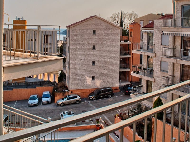 Pogled sa balkona stana u Petrovcu, Crna Gora: moderne stambene zgrade, parking i morski pejzaž na horizontu.