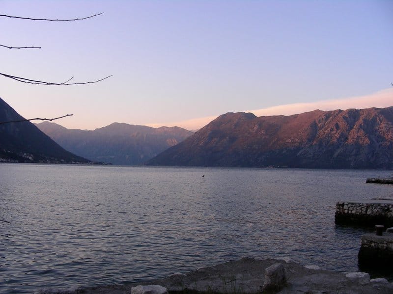 Vue panoramique sur la baie de Kotor et les montagnes depuis une maison à Dobrota, Monténégro : paysage marin avec un quai en pierre au coucher du soleil.