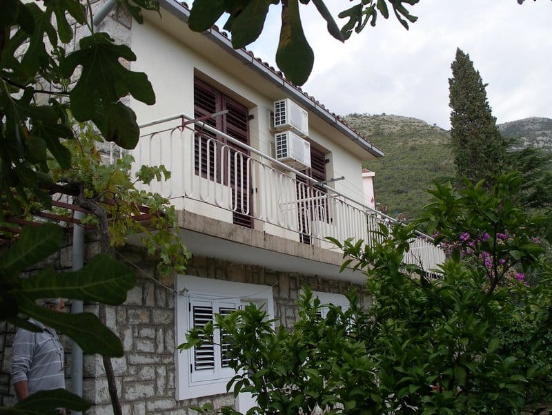 Facade of a stone house in Petrovac, Montenegro, with a long balcony, air conditioners, and a green garden against a mountain backdrop.