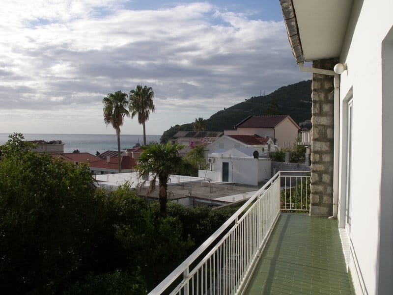 View from the balcony of a house in Petrovac, Montenegro: a panorama of the sea, palm trees, mountains, and the city’s tiled roofs.