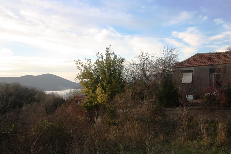 Dve kamene kuće sa crepnim krovom i pogledom na more u Krašićima, Crna Gora, okružene baštom i brežuljkastim pejzažem.