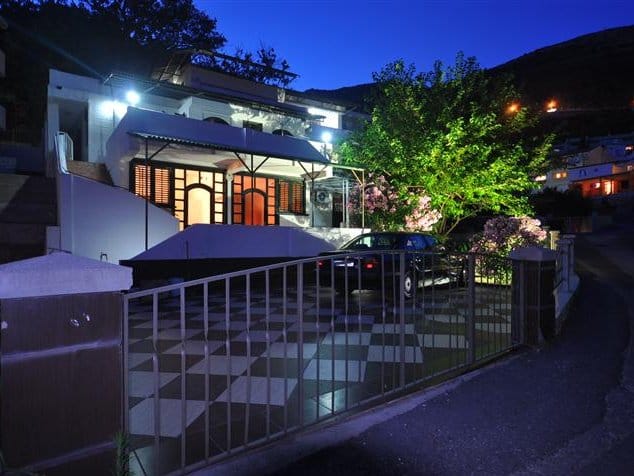 Two-story house in Petrovac, Montenegro: night view of the facade, terrace, parking area with checkerboard paving, and garden.