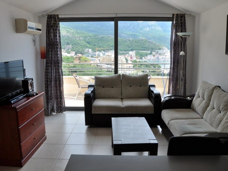 Salon avec vue panoramique sur la mer et les montagnes dans un appartement à Bečići, Monténégro : mobilier moelleux, terrasse et coin TV.