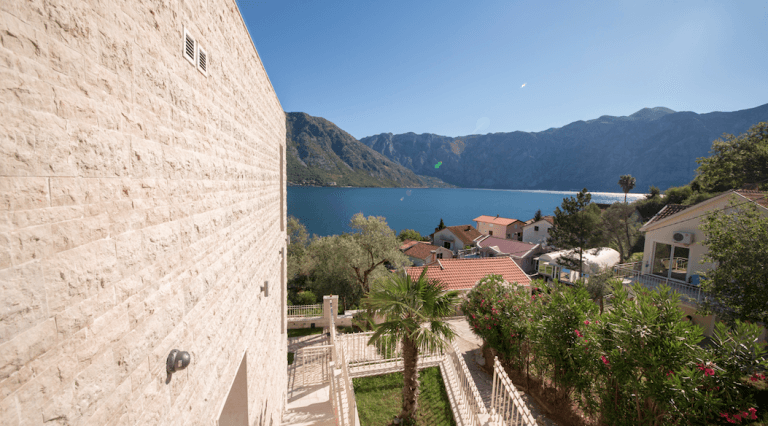 Blick auf die Bucht von Kotor und die Berge von der Terrasse eines Hauses in Stoliv, Montenegro: Steinfassade und gepflegter Garten mit Palme.