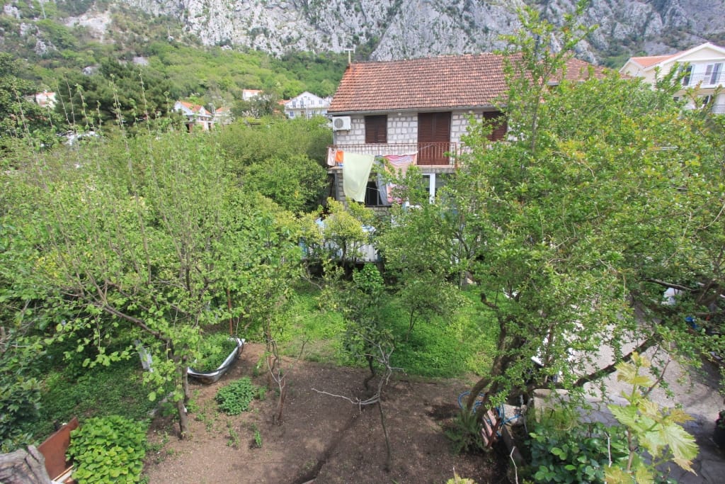 Maison en pierre de deux étages avec jardin à Dobrota, Monténégro : vue sur la façade, terrain verdoyant et paysage montagneux en arrière-plan.
