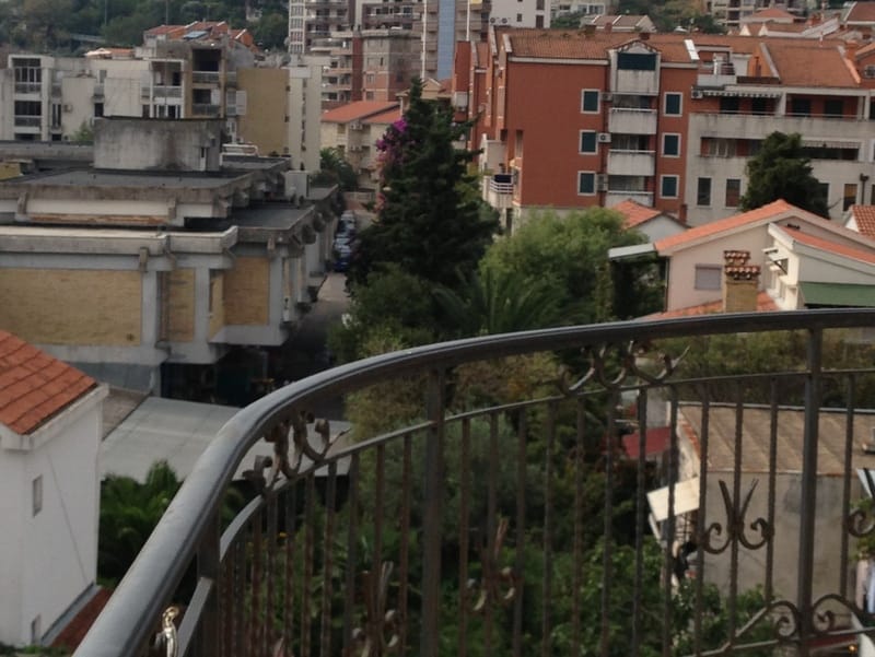 Pogled s balkona na gradske kuće i zelenilo u Budvi, Crna Gora: kovana ograda i panorama stambenog kvarta.