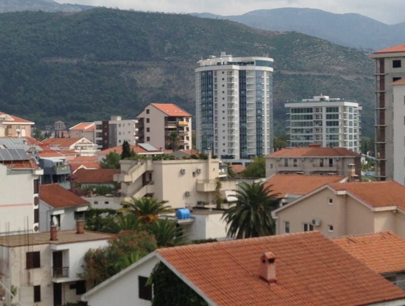 Panoramski pogled na stambene zgrade i moderne nebodere u gradu Budvi, Crna Gora, sa planinama u pozadini.