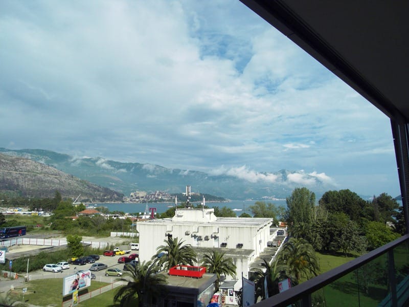 Pogled sa balkona stana u Budvi, Crna Gora: panorama planina, mora i gradske infrastrukture sa palmama.