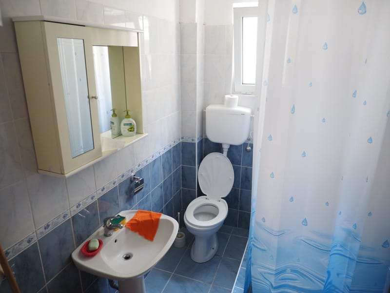 Badezimmer in einem Haus in Bar, Montenegro: helle Fliesen, Waschbecken, Toilette und Duschbereich mit Vorhang.