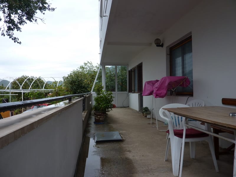 Prostran balkon kuće u Sutomoru, Crna Gora: drveni sto, plastične stolice i pogled na zelenu baštu.