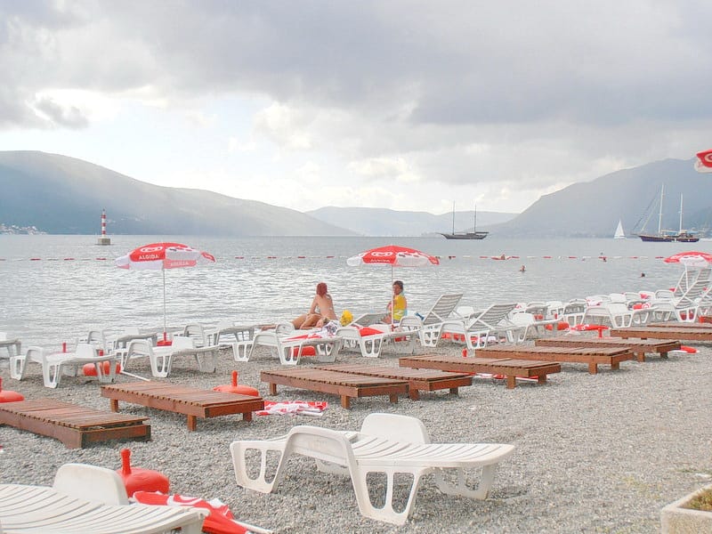Plaža sa ležaljkama i suncobranima uz more u Tivtu, Crna Gora: pogled na zaliv, planine i jahte pored hotela.