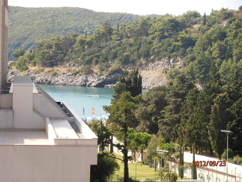 Pogled sa balkona stana u Bečićima, Crna Gora: panorama morske uvale, plaže i zelenog šumovitog planinskog obronka.