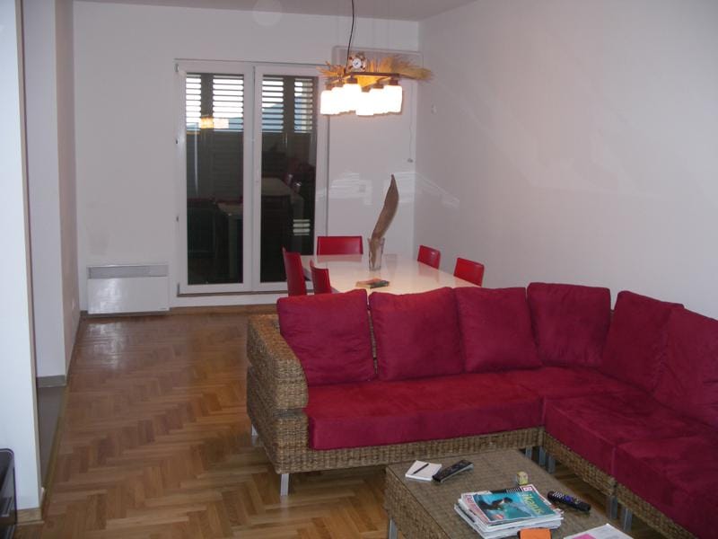 Salon avec canapé en osier et coin repas dans un appartement à Herceg Novi, Monténégro : parquet, intérieur lumineux.
