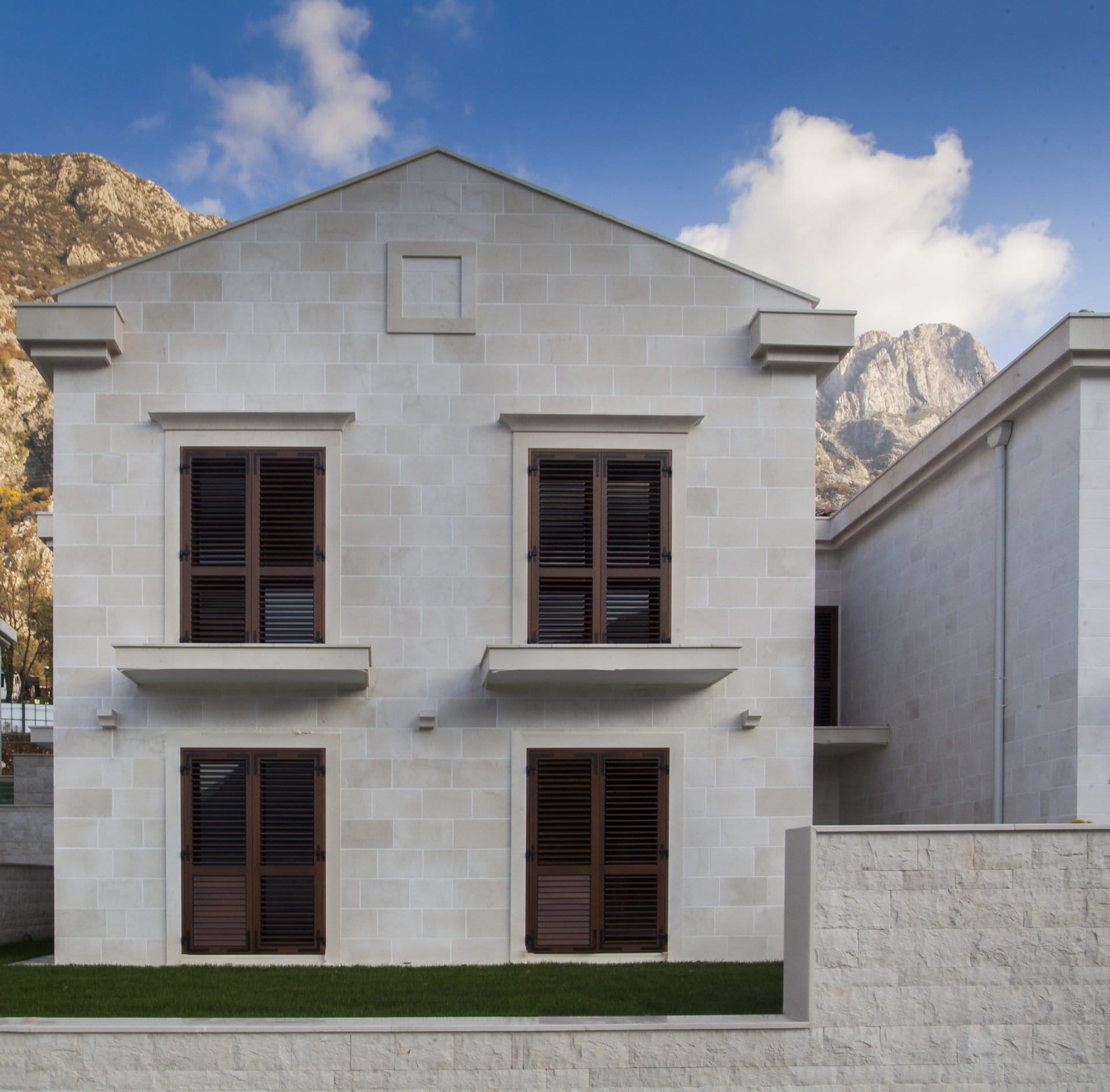 Fassade einer modernen Villa aus hellem Stein mit braunen Fensterläden in Dobrota, Montenegro, vor einer Bergkulisse.