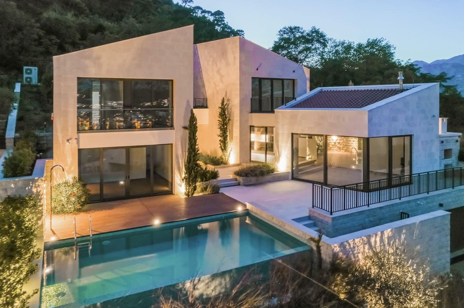 Современная вилла в Прчани, Черногория: двухэтажный дом с панорамными окнами, террасой и открытым бассейном вечером.
