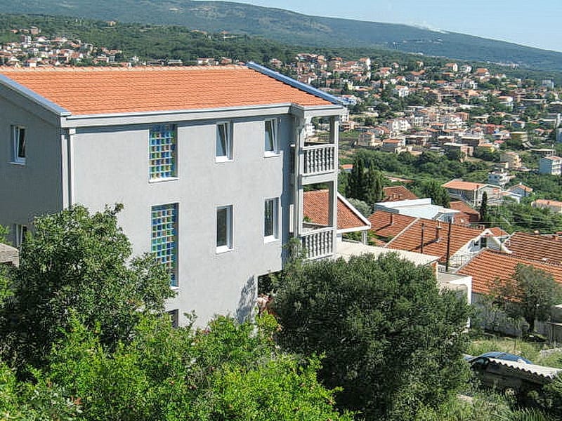 Трехэтажный дом с черепичной крышей и балконами в Добрых Водах, Черногория, на фоне горного пейзажа и города.