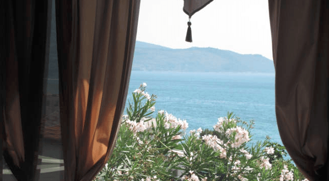 Pogled na more i planine iz prozora stana u Igalu, Crna Gora, uokviren braon zavesama sa rascvetalim oleandrom.