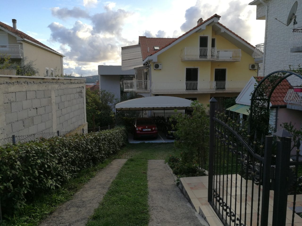 Dvospratna kuća u Tivtu, Crna Gora, sa privatnim parkingom pod nadstrešnicom, zelenim dvorištem i kovanom kapijom.