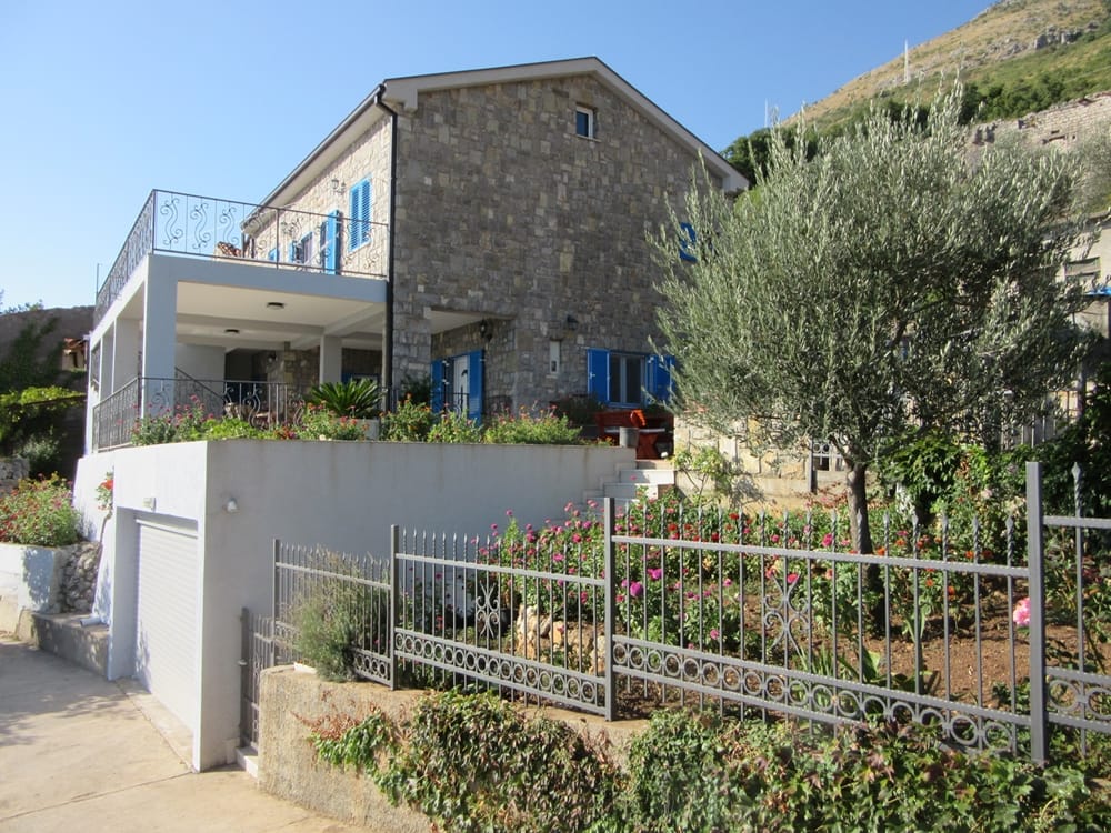 Каменная вилла в Режевичи, Черногория: фасад с террасой, гаражом, цветущим садом и оливковым деревом на участке.