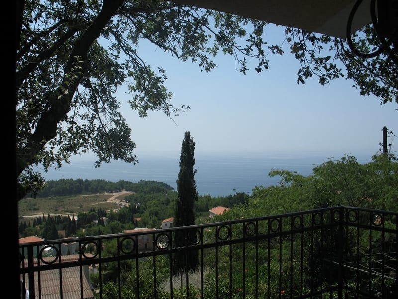 Widok na morze i zielone wzgórza z tarasu domu w Sutomore, Czarnogóra, z kutym ogrodzeniem i koronami drzew.