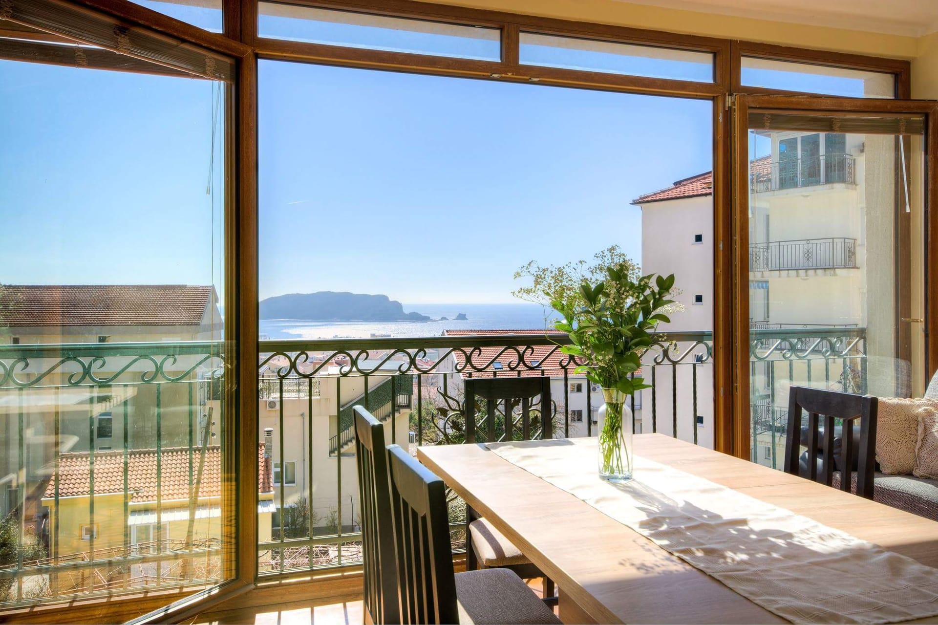 Blick auf das Meer und die Insel vom Essbereich eines Apartments in Budva, Montenegro: Panoramafenster, Holztisch und Balkon.