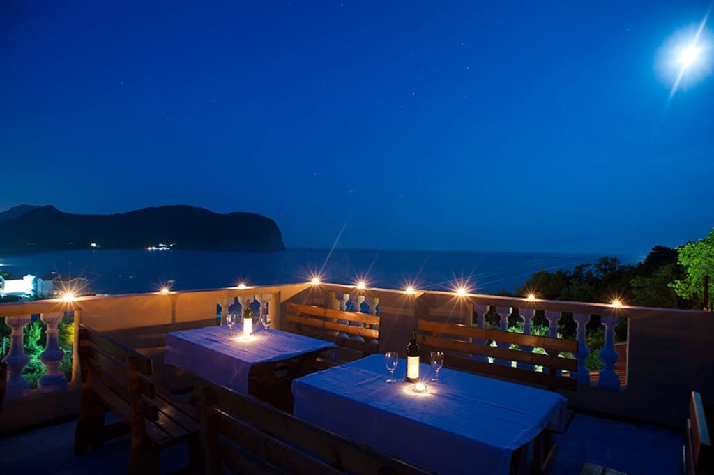 Hotel terrace in Buljarica, Montenegro: wooden tables with place settings and a panoramic view of the sea and mountains at night.