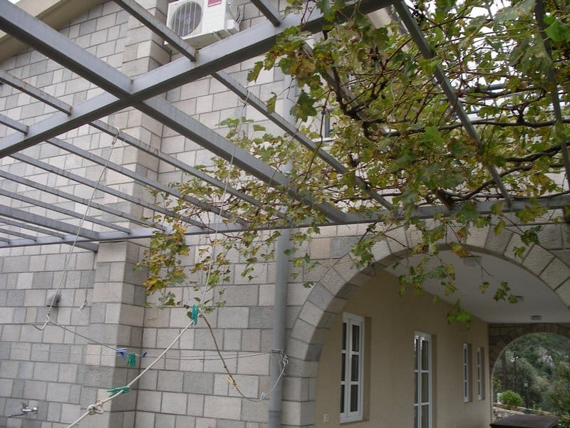 Терраса дома в Режевичи, Черногория: каменная кладка, арочный проход и навес с виноградной лозой над зоной отдыха.