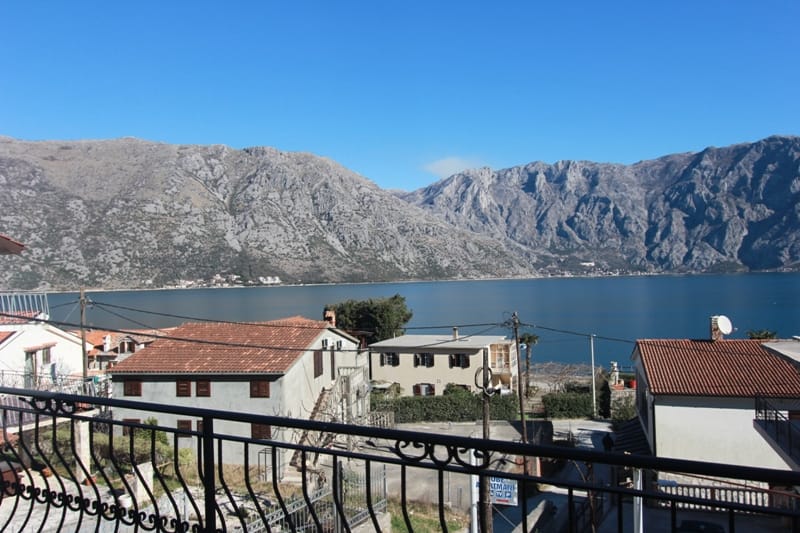 Vue depuis le balcon d'une maison à Stoliv, Monténégro : panorama de la baie de Kotor et des montagnes, toits en tuiles et garde-corps en fer forgé.