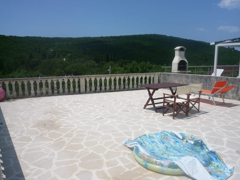 Villa terrace in Bigovo, Montenegro: stone floor, outdoor furniture, barbecue, and panoramic view of green mountains.