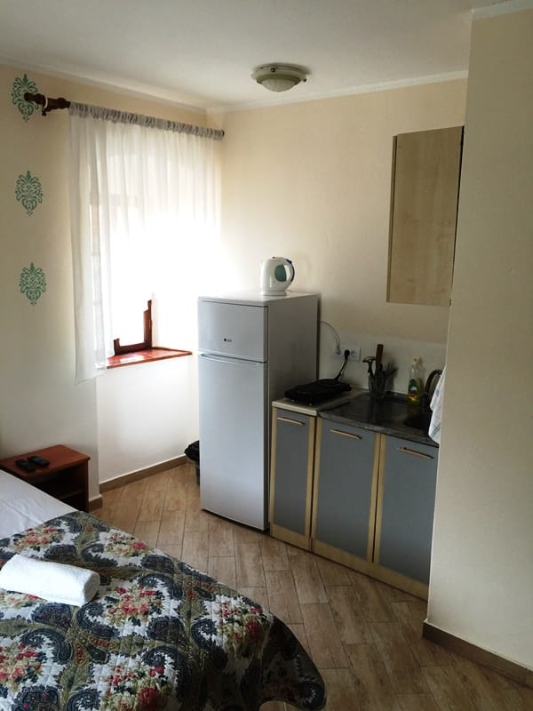 Компактная кухня в номере отеля в Дженовичи, Черногория: холодильник, плита, мойка и кровать с узорчатым покрывалом.