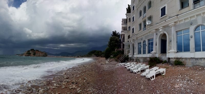 Restaurant en bord de mer à Sveti Stefan, Monténégro : vue sur la plage, chaises longues et l'île de Sveti Stefan sous un ciel nuageux.
