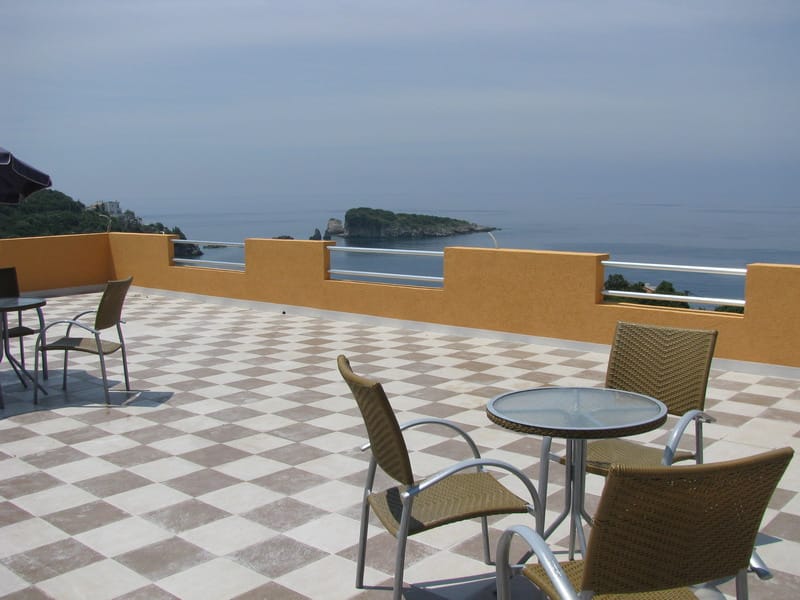 Terrasse avec fauteuils en osier et vue sur la mer dans une maison à Utjeha, Monténégro : carrelage en damier et paysage marin panoramique.