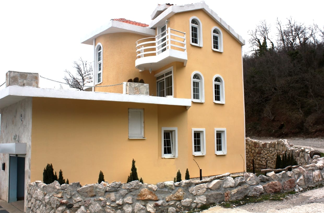 Трехэтажная вилла в Марковичи, Черногория: желтый фасад, арочные окна, балконы и каменная ограда на фоне леса.