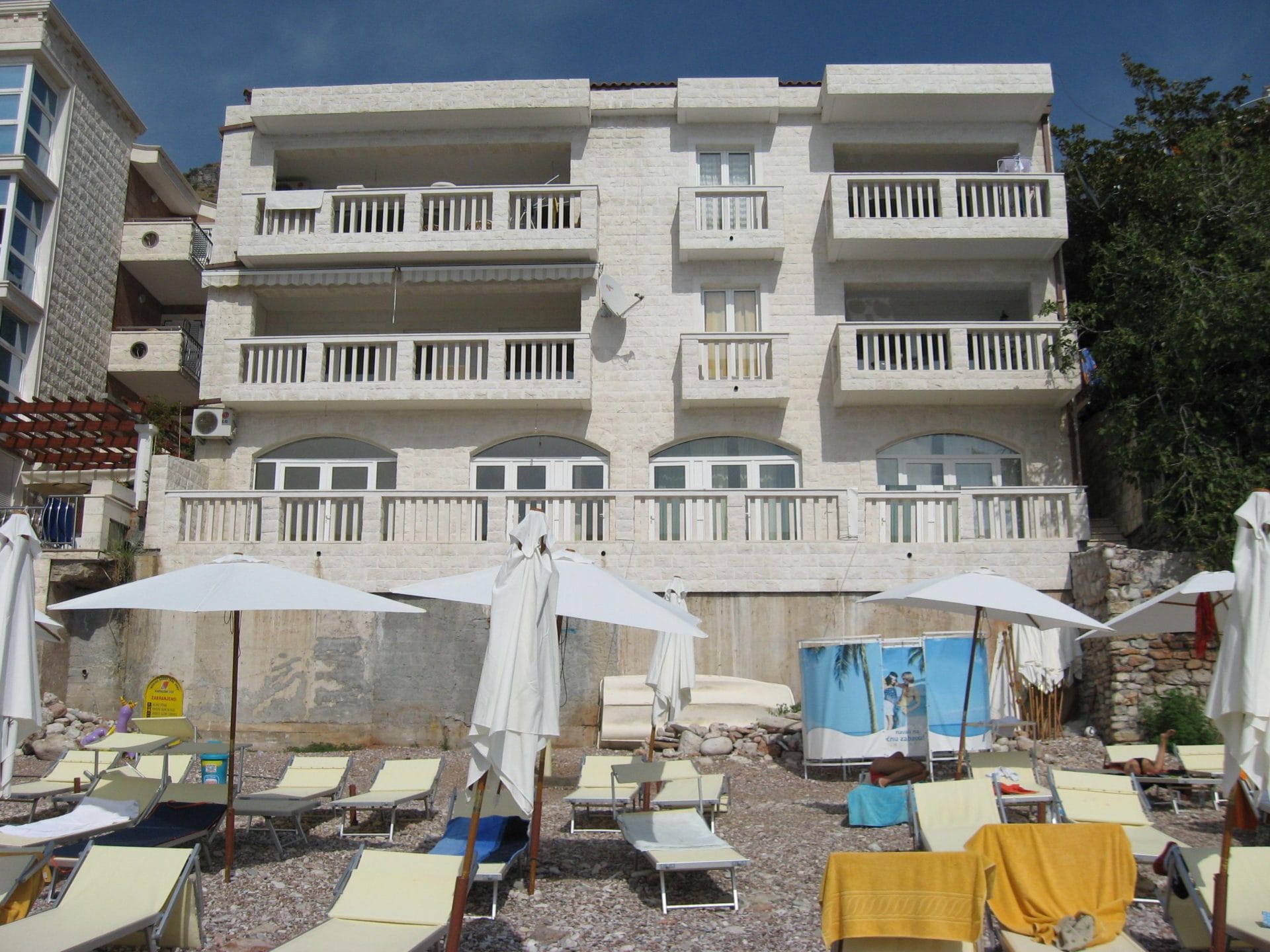 Maison de trois étages avec balcons blancs sur la plage de Sveti Stefan, au Monténégro, avec chaises longues et parasols au bord de la mer.
