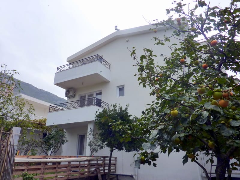 Белый двухэтажный дом с балконами и садом в Сутоморе, Черногория: вид на фасад с фруктовыми деревьями и террасой.