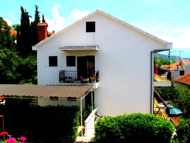 Белый двухэтажный дом с балконами и террасой в Герцег-Нови, Черногория, окруженный зеленью и деревьями.