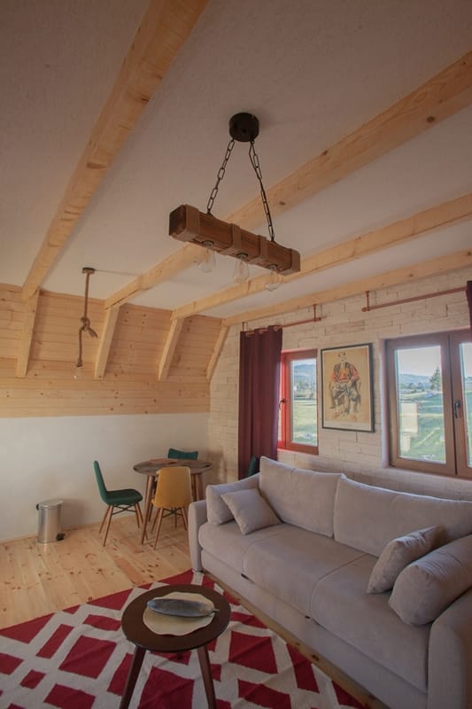 Salon w stylu rustykalnym w domu w Žabljaku, Czarnogóra: drewniane belki, jasna sofa, jadalnia i widok na góry.