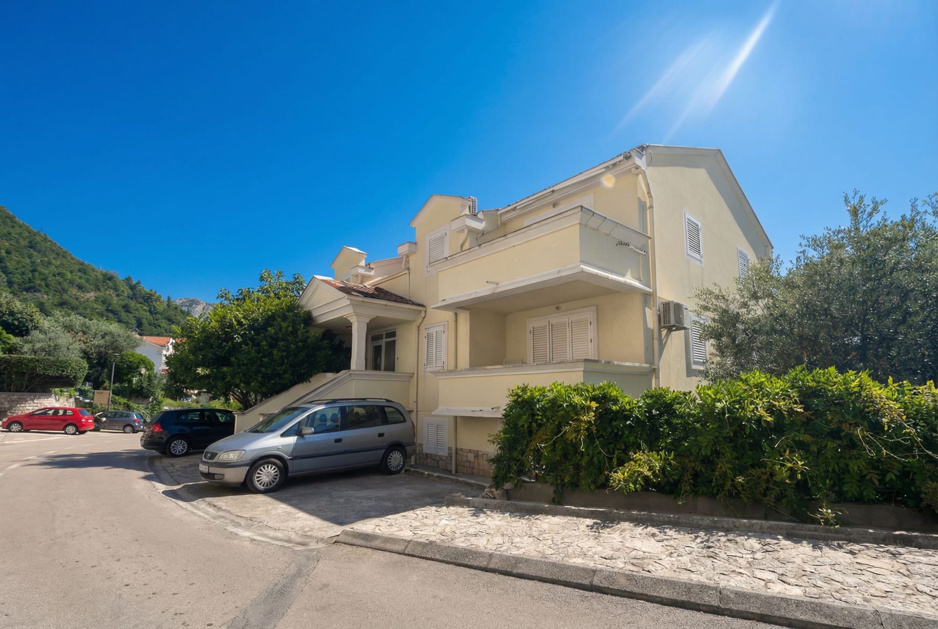 Two-story house in Sveti Stefan, Montenegro: facade with balconies, parking, and greenery around the building.