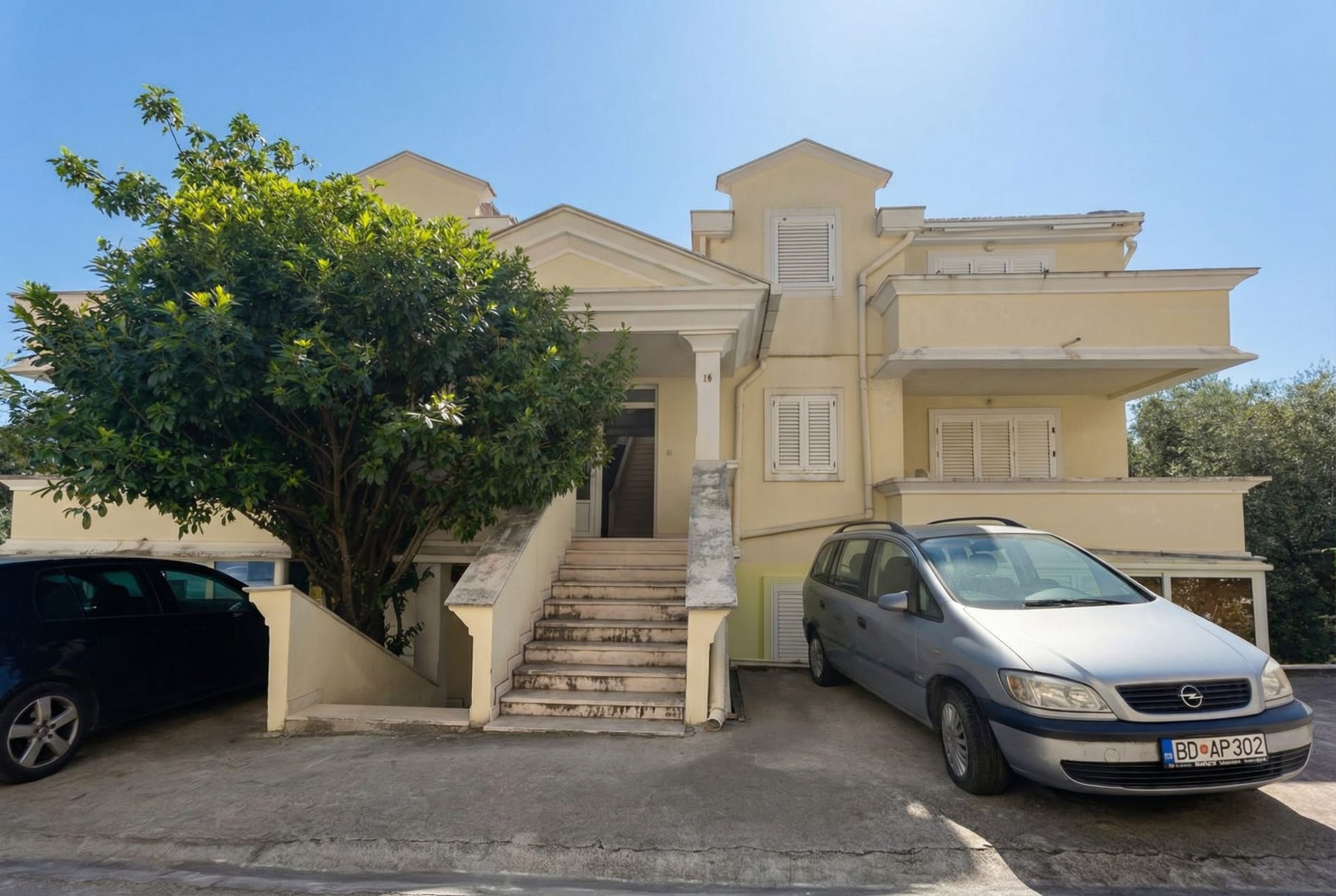 Facade of a two-story house in Sveti Stefan, Montenegro: light walls, white shutters, entrance stairs, and parking by the house.