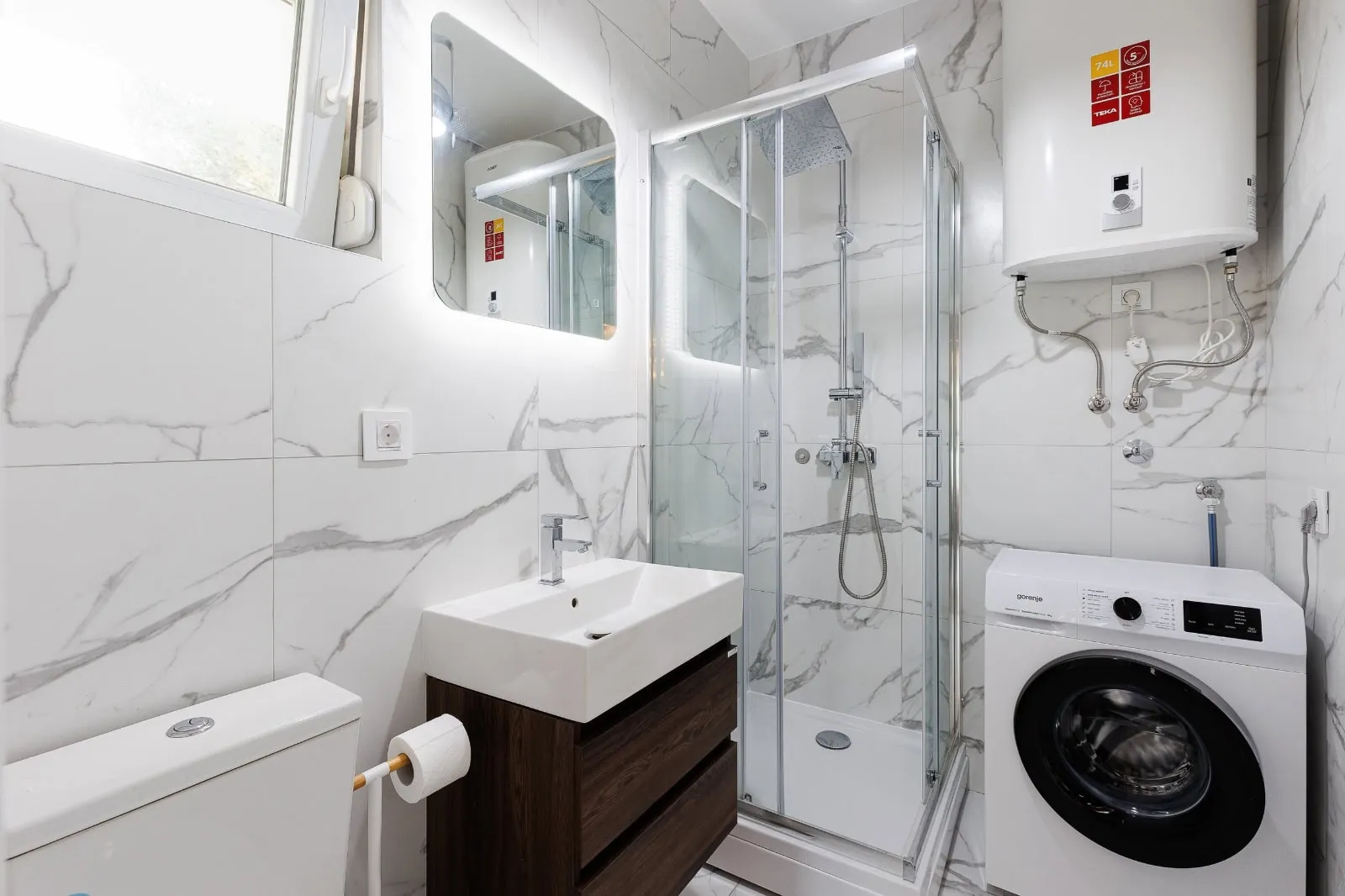Salle de bain avec cabine de douche en verre, vasque sur meuble bois, toilette, machine à laver et chauffe-eau mural.