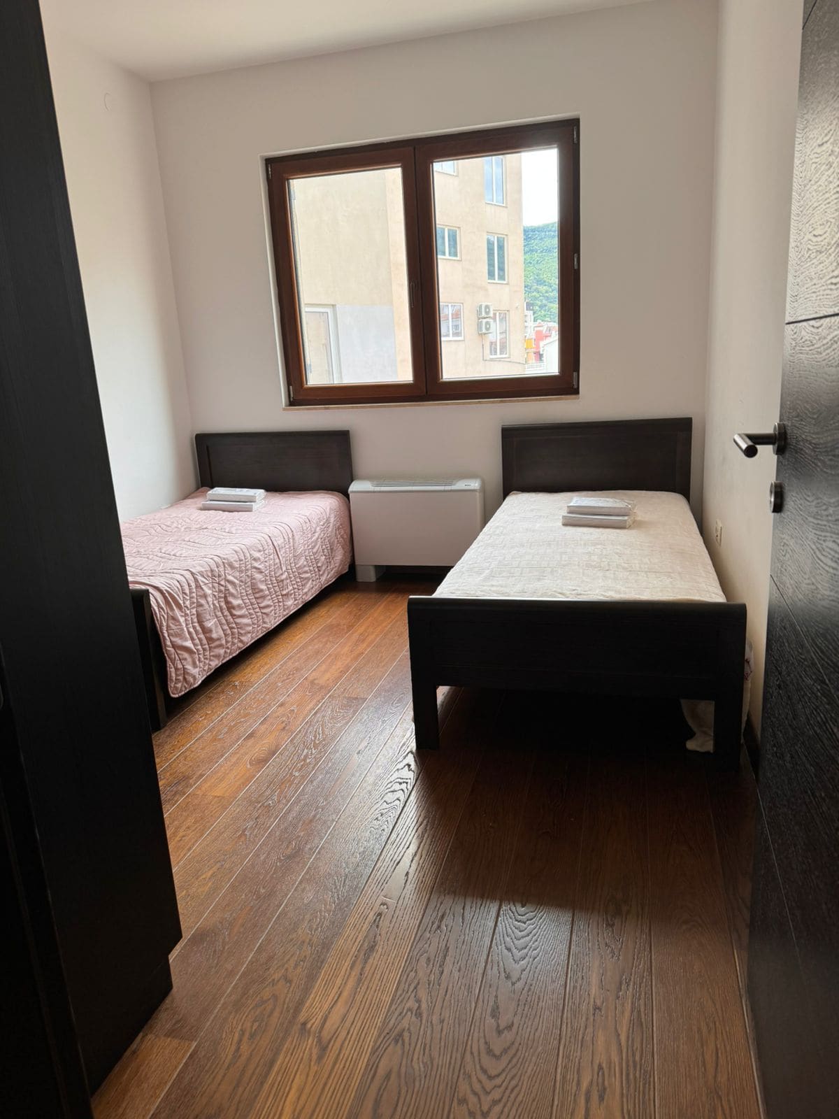 Schlafzimmer mit zwei Betten, dunklen Möbeln und Parkett in einer Wohnung in Budva, Montenegro. Blick auf die Stadt aus dem Fenster.