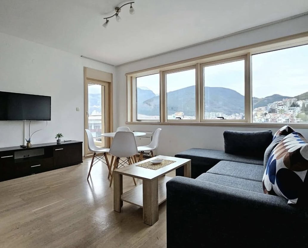 Bright living room in an apartment in Bečići, Montenegro: gray sofa, dining area, and panoramic mountain views from the windows.