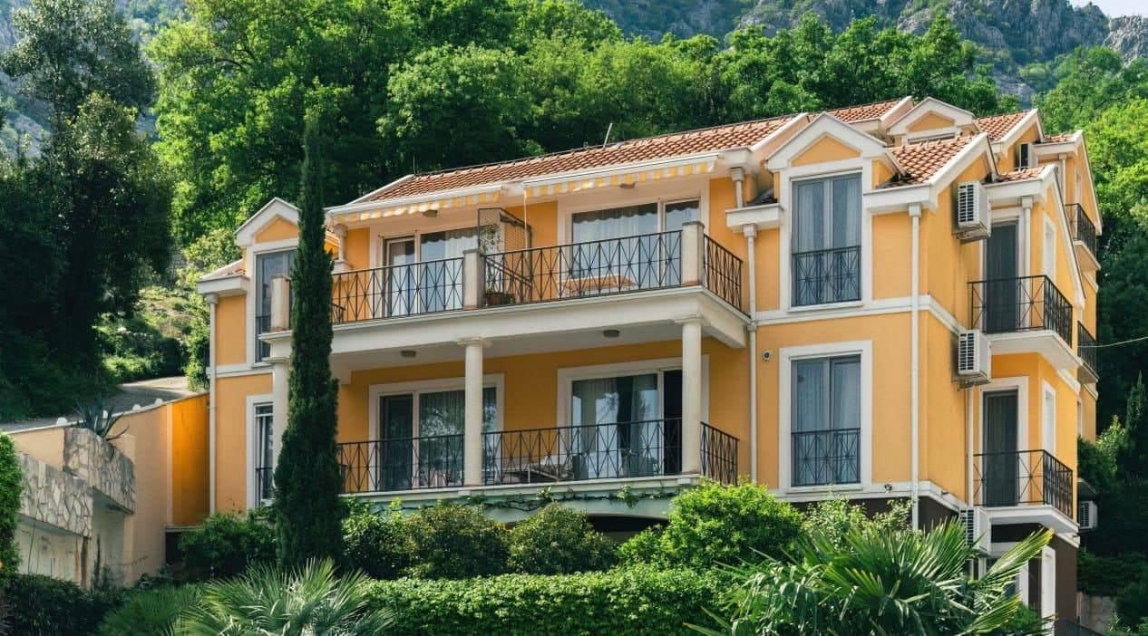 Trospratna žuta kuća sa balkonima i krovom od crepa u Orahovcu, Crna Gora, okružena zelenim drvećem.